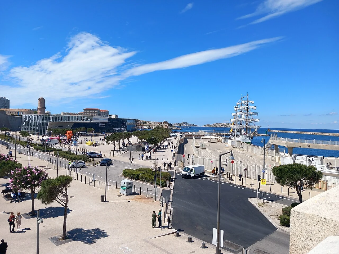 Esplanade et quais près du Mucem, un grand voilier au loin sous un ciel bleu.